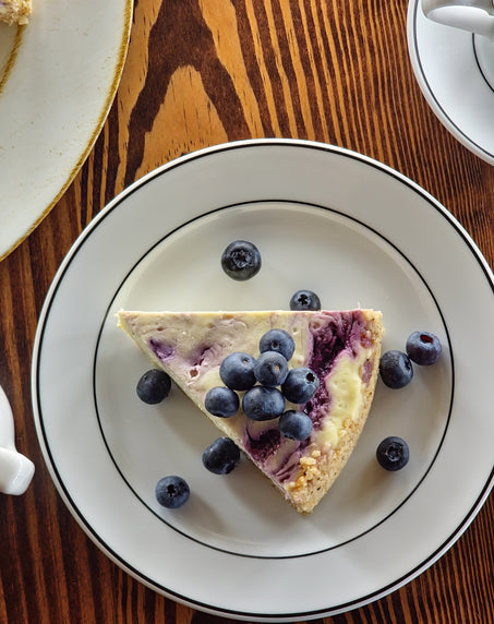 A slice of blueberry cheesecake topped with fresh blueberries on a white plate, accompanied by a cup of coffee, a small pitcher of cream, and a bowl of sugar on a wooden table.