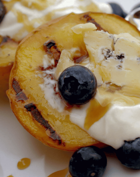 Grilled peach halves topped with whipped cream, blue cheese, blueberries, and drizzled with honey on a white plate.
