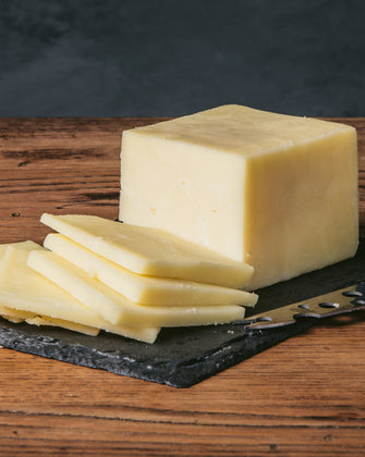 A block of Jefferson Cheddar cheese, partially sliced, placed on a slate board with a cheese knife beside it.
