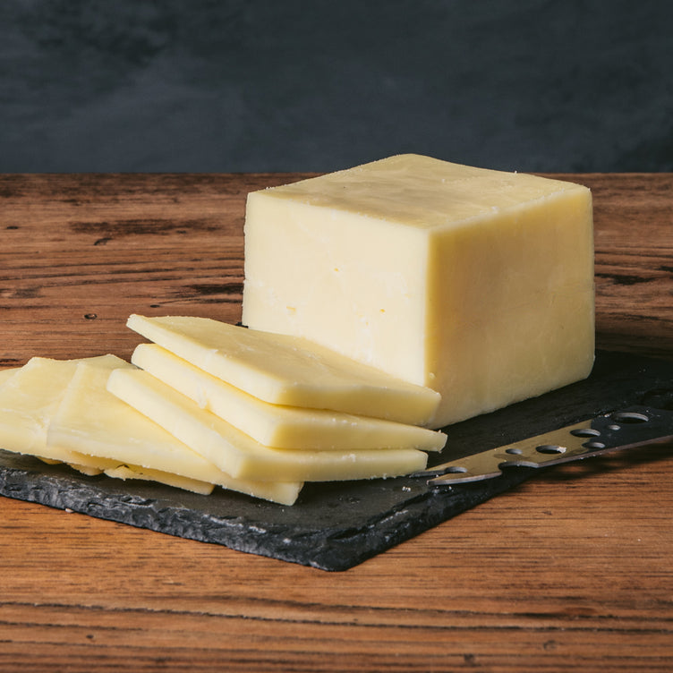 A block of Jefferson Cheddar cheese, partially sliced, placed on a slate board with a cheese knife beside it.
