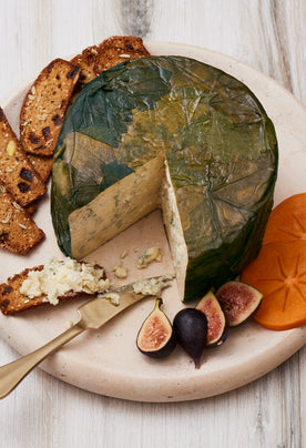 A round cheese wrapped in green leaves on a wooden board, served with fig slices, persimmon slices, and nut-studded crackers with cheese spread.