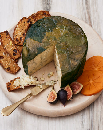 A round cheese wrapped in green leaves on a wooden board, served with fig slices, persimmon slices, and nut-studded crackers with cheese spread.