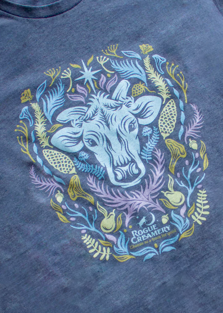 A heather blue t-shirt featuring a cow’s head surrounded by colorful Southern Oregon flora, with “Rogue Creamery” printed below.
