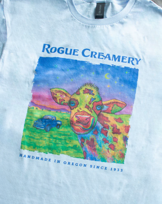 A blue t-shirt featuring a vibrant, multicolored cow illustration with a vintage truck and night sky, “Rogue Creamery” and “handmade in Oregon since 1933” printed on the front.