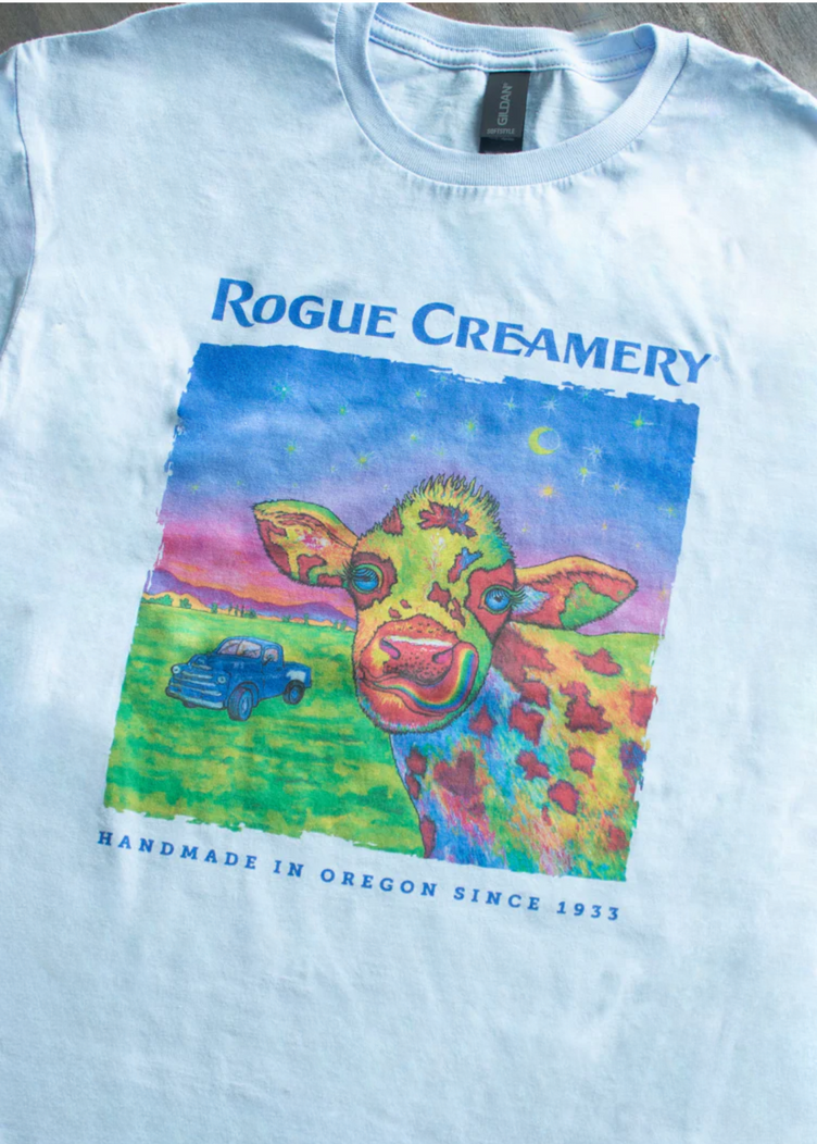 A blue t-shirt featuring a vibrant, multicolored cow illustration with a vintage truck and night sky, “Rogue Creamery” and “handmade in Oregon since 1933” printed on the front.